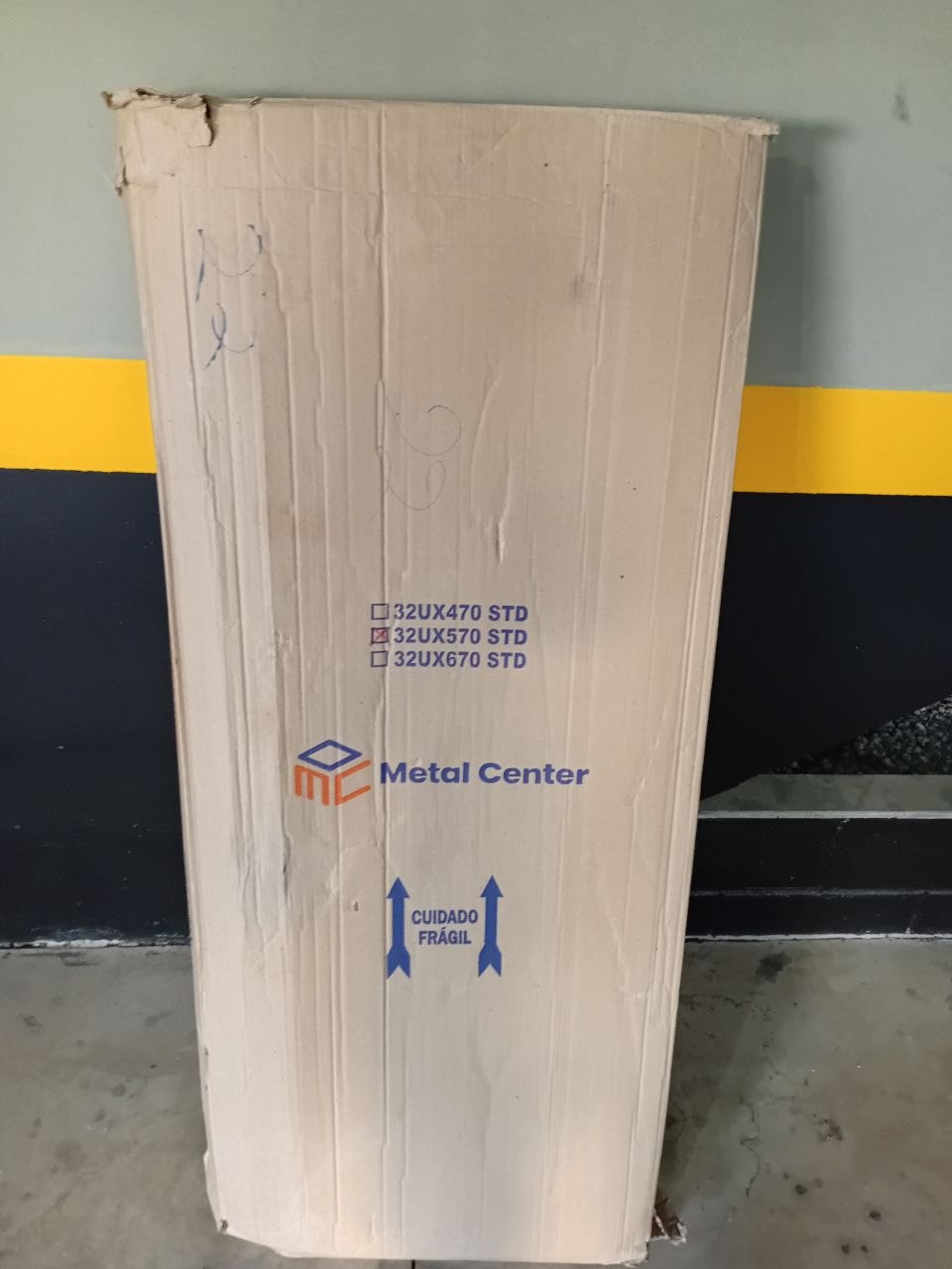 Embalagem de papelão original (Metal Center) — canto superior já rasgado/frágil, com CUIDADO FRÁGIL e NÃO PISE impressos.