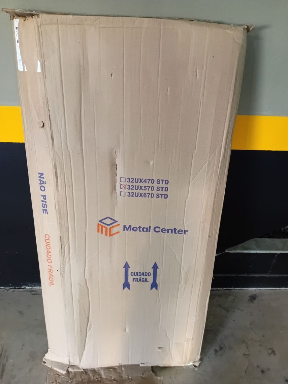 Embalagem de papelão original (Metal Center) — canto superior já rasgado/frágil, com CUIDADO FRÁGIL e NÃO PISE impressos.